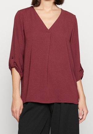 Femme portant un chemisier ample bordeaux à col en V avec des manches retroussées aux trois quarts et un pantalon large noir sur un fond uni.