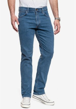 Homme portant un jean slim bleu, un t-shirt blanc, des baskets blanches et une montre-bracelet argentée, debout devant un fond blanc.