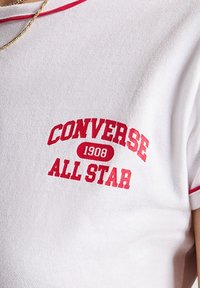 Camiseta blanca de manga corta con ribete rojo que presenta el logo "CONVERSE 1908 ALL STAR" en letras rojas negritas en la parte frontal. Textura de tela suave.