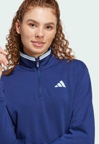 Marinblå sweatshirt med hög krage, halv dragkedja och vitt logotypaccent. Slät textur med en kontrasterande ljus rand vid kragen.
