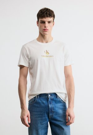Młody mężczyzna w białym t-shircie Calvin Klein Jeans z złotym logo oraz niebieskich dżinsach, stoi na tle jednolitego białego tła.
