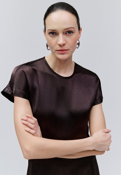 Femme aux yeux bleus et cheveux foncés plaqués en arrière, portant des boucles d'oreilles en argent en forme de cercle et un haut en satin marron foncé à manches courtes, bras croisés.