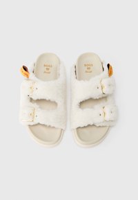Sandales blanches duveteuses avec deux boucles dorées réglables, semelles en beige et une étiquette de marque. Texture douce avec un design ouvert.