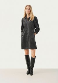 Schwarzes Cordkleid mit langen Ärmeln, Frontreißverschluss, seitlichen Taschen und tailliertem Schnitt, kombiniert mit kniehohen schwarzen Lederstiefeln.