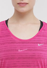Rosa träningsskjorta med ljusa horisontella ränder, rund halsringning, prydd med grå Nike-logotyp på bröstet. Strukturerad, andningsbar tyg.