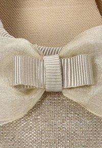 Zapato de tela beige con una superficie texturizada, que presenta un gran lazo de color crema claro con bordes ondulados y un patrón de franjas verticales en el centro.