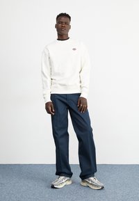 Sudadera blanca con logo, pantalones azules y zapatillas grises. La tela parece suave; las zapatillas tienen un diseño texturizado y en capas.