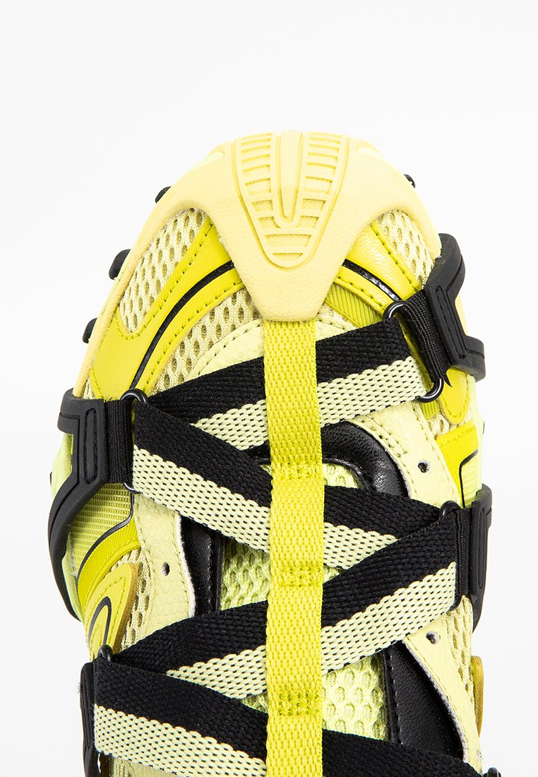 Gelbe und schwarze Sportschuhe aus Netzstoff mit kreuzförmig angeordneten schwarzen und gelben Riemen, die durch Kunststoffschnallen gesichert sind.