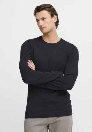 Zwart gebreid sweater met een textuurpatroon, ronde halslijn en ribbelboorden. Draagt over lichtbeige broeken tegen een effen achtergrond.