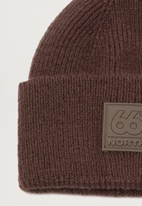 Ruskea neulottu pipo, jossa on paksu ribbitekstuuri ja taitettu reuna. Pipossa on neliskulmainen nahkaleima, jossa on kaiverrettuna "66° NORTH".