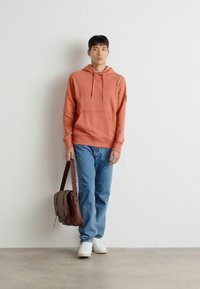 Felpa con cappuccio arancione chiaro, dotata di tasca a marsupio, abbinata a jeans blu. Il modello tiene una borsa a tracolla marrone e si trova in piedi contro un muro semplice.