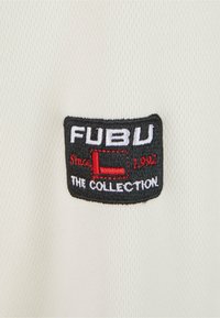Hvit stoff T-skjorte med en textured overflate, med et svart brodert merkelapp som står "FUBU THE COLLECTION Siden 1992" i hvitt og rødt.