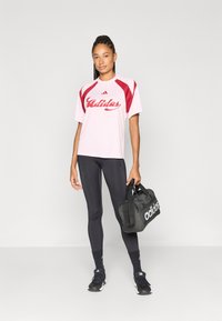 Adidas roze en rode shirt met korte mouwen en wit logo, zwarte legging, zwarte sporttas en zwarte sportschoenen. Glad materiaal, nauwsluitend ontwerp.