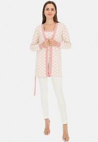 Długo rękawowy kardigan w jasnoróżowym kolorze z geometrycznymi wzorami, z kontrastującą różową lamówką i związanym w pasie. Nosi się go na różowej bluzce i białych spodniach.