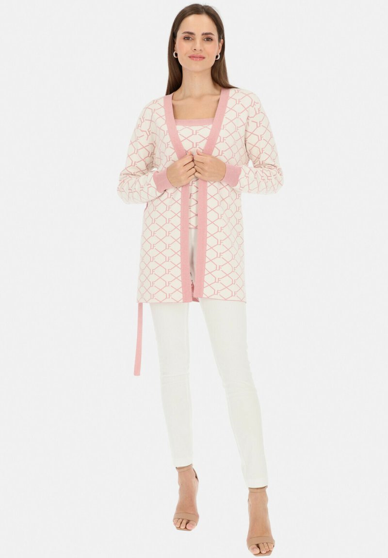 Długo rękawowy kardigan w jasnoróżowym kolorze z geometrycznymi wzorami, z kontrastującą różową lamówką i związanym w pasie. Nosi się go na różowej bluzce i białych spodniach.