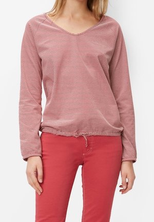 Žena nosící červenobílé proužkované tričko s dlouhým rukávem a výstřihem do V, s nastavovacím stahováním na spodním okraji, v kombinaci s jednobarevnými červenými úzkými kalhotami.