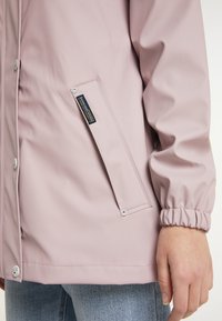 Veste imperméable rose avec des boutons-pression, des poignets élastiques et une poche latérale avec une étiquette logo. Fabriquée en matériau synthétique lisse.