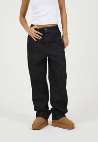 Jeans de mezclilla negros de pierna ancha con costuras en contraste, cinco bolsillos y un ajuste de cintura alta. Usados con una camiseta blanca ajustada y zapatos marrones sin cordones.