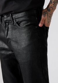 Jeans de denim negro con un acabado elegante, diseño de cinco bolsillos y acentos de botón metálico. Tejido texturizado con un sutil brillo.