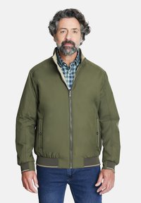 Chaqueta bomber en verde oliva con cierre de cremallera, puños y dobladillo de canalé. Los bolsillos tienen sutiles detalles metálicos. Estilizada sobre una camisa de cuadros.