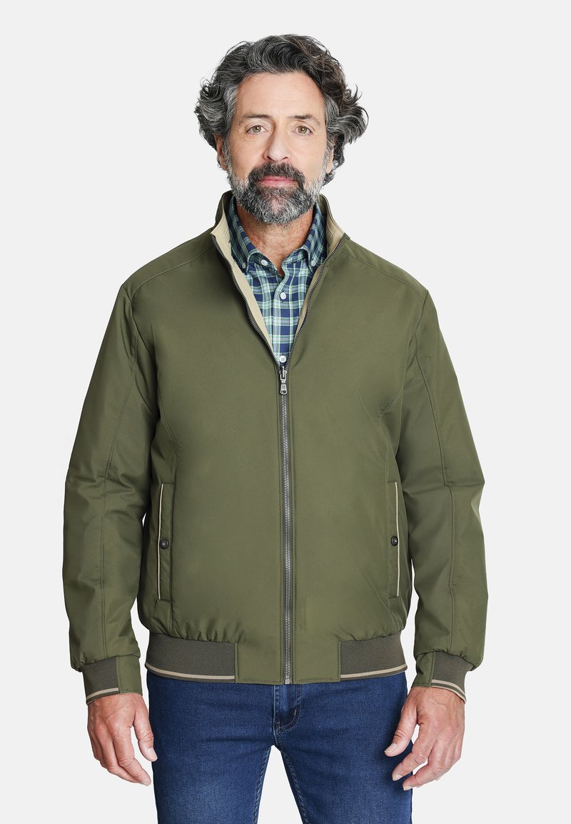Chaqueta bomber en verde oliva con cierre de cremallera, puños y dobladillo de canalé. Los bolsillos tienen sutiles detalles metálicos. Estilizada sobre una camisa de cuadros.