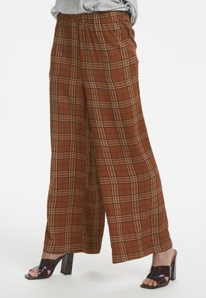 Szerokie brązowe spodnie w kratę z elastycznym paskiem, noszone z czarnymi sandałami na obcasie typu block z otwartym noskiem na białym tle.
