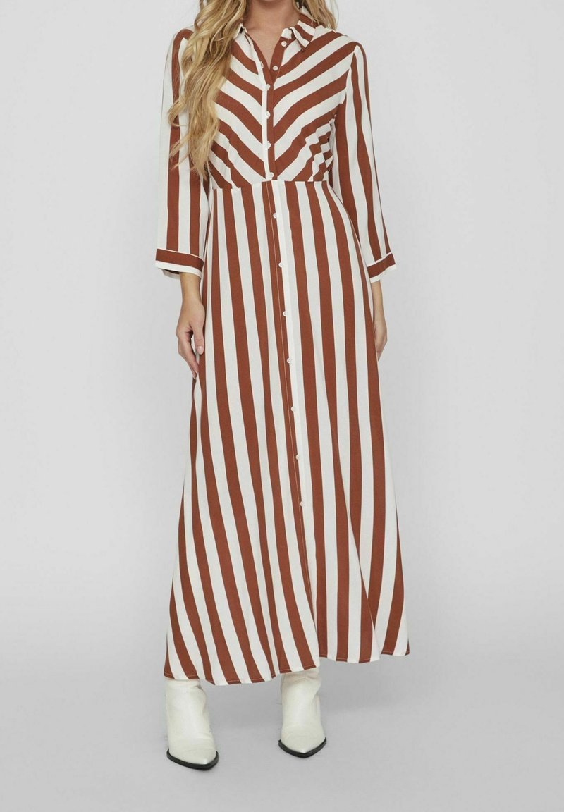 Femme portant une longue robe chemise à rayures verticales marron et blanches, manches longues, et des bottines blanches.