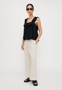 Top nero senza maniche con scollatura arricciata, abbinato a pantaloni beige a gamba larga e sandali neri con tacco a blocco, stilizzato con occhiali da sole.