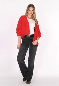 Rode, geweven cardigan met lange mouwen, open voorkant, gedragen over een witte top en zwarte high-waisted jeans. Zwarte laarzen maken de outfit compleet.