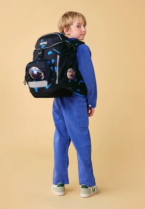 Child in blue jumpsuit wearing a large black and blue backpack with car and skateboard patches, standing against a beige background.