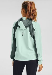 Veste vert menthe avec une capuche vert foncé, présentant une coupe slim, une texture lisse et des lignes d'accent horizontales dans le dos.