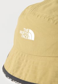 Μπεζ καπέλο τύπου bucket με λευκό λογότυπο The North Face στο μπροστινό μέρος και μαύρο περίγραμμα με λευκό κείμενο κατά μήκος της άκρης του γείσου.
