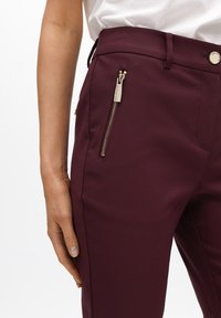 Pantalones ajustados burdeos hechos de una tela suave, con un bolsillo con cremallera dorada y un cierre de botón en la cintura para un detalle adicional.