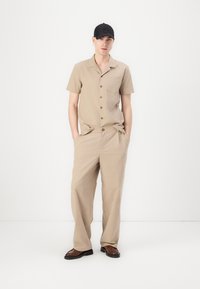 Camisa beige de manga corta con botones, combinada con pantalones beiges de corte holgado. El conjunto cuenta con una tela texturizada y un bolsillo en el pecho.