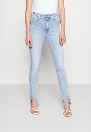 Personne portant un jean skinny taille haute bleu clair avec des fentes à l’avant des chevilles et des sandales blanches à brides sur fond uni.