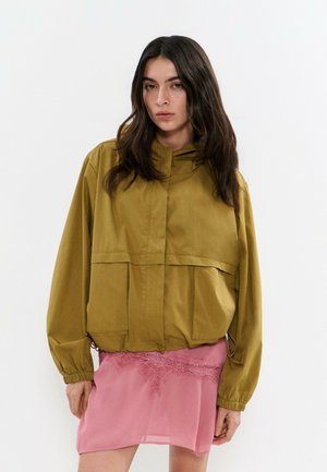 Mujer con cabello largo y oscuro que lleva una chaqueta verde oliva con grandes bolsillos y puños elásticos, combinada con una falda corta rosa con detalles de encaje.