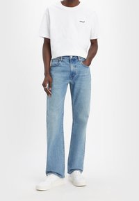 Camiseta blanca de algodón con logo negro, combinada con jeans rectos de color azul claro. Zapatillas blancas completan el atuendo. Diseño simple y casual.