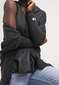 Osoba oblečená v černém bezramenném topu, černém dlouhém kardiganu částečně spadlém z jednoho ramene, a světle modrých džínách, sedící se zkříženýma rukama.