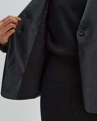 Schwarze Lederblazer mit Knopfverschluss, Innenfutter sichtbar. Schwarzer Strickpullover darunter, der ein figurschmeichelndes Design zeigt.