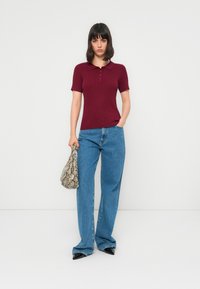 Donna che indossa una polo a maniche corte bordeaux, jeans a gamba larga blu, scarpe nere con punta, e tiene una borsa con motivo in pelle di serpente.