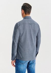 Mężczyzna w szarej koszulce z długimi rękawami i niebieskich dżinsach, stojący plecami do aparatu na białym tle.