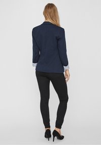 Blazer blu navy con un design aderente, dettagli a righe leggere sui polsini e pantaloni neri eleganti abbinati a tacchi alti neri. Tessuto dalla consistenza liscia.