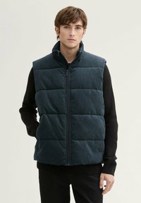 Dunkelgrüne gepolsterte Weste aus Cordstoff, mit hohem Kragen, Frontreißverschluss und gestepptem Design. Getragen über einem schwarzen strukturierten Pullover.
