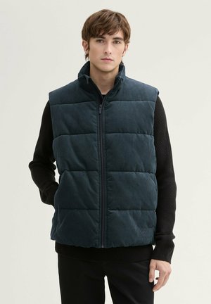 Dark green padded vest made of corduroy, featuring a high collar, front zipper, and quilted design. Worn over a black textured sweater.