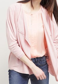 Cardigan rosa chiaro con un design fluido, texture morbida e apertura frontale, indossato sopra una blusa pesco chiara con dettagli plissettati.