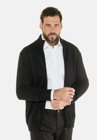 Schwarze, mit Reißverschluss versehene Strickjacke mit gerippten Bündchen und Kragen, getragen über einem weißen Hemd. Glatte Textur, taillierter Schnitt, minimale Details.