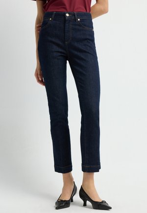 Donkerblauwe, high-waisted jeans met een aansluitende silhouette, opgerolde zoom en gouden stikdetails. Gecombineerd met zwarte, puntige hakken.