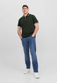 Polo shirt verde scuro con colletto a righe bianche e gialle, abbinato a jeans azzurri chiari e sneakers bianche. Figura maschile in piedi.