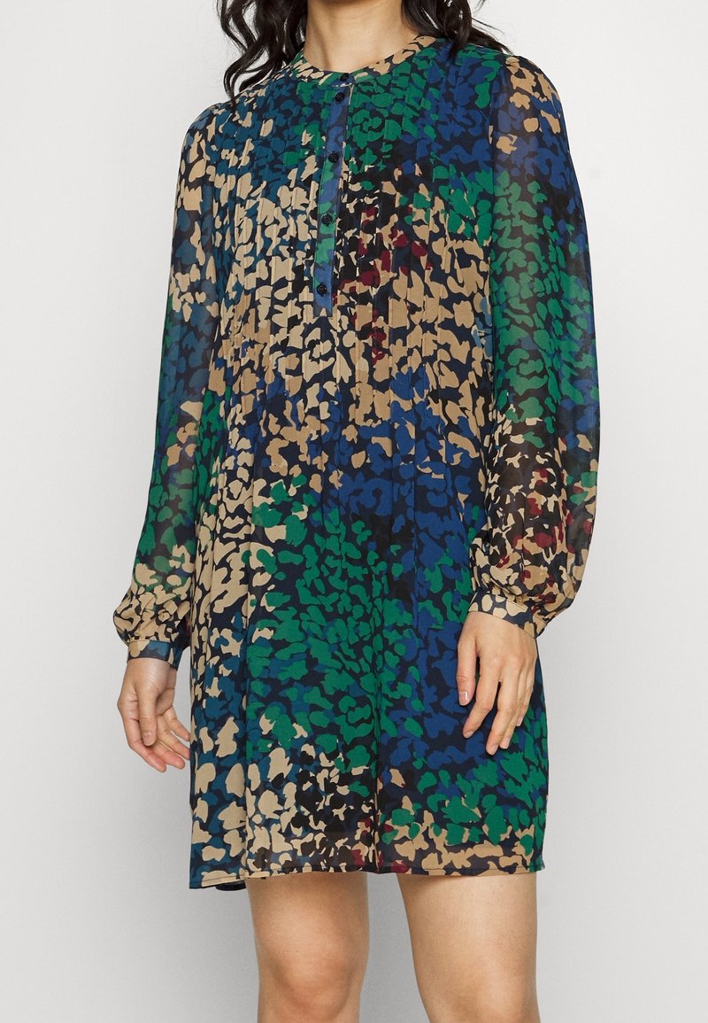 Femme portant une robe à manches longues boutonnée avec un motif floral abstrait bleu, vert, beige et noir.