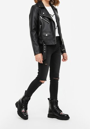 Donna che indossa una giacca da motociclista in finta pelle nera, jeans skinny neri strappati, camicia bianca con stampa a cuore e stivali anfibi neri con cuori rossi.